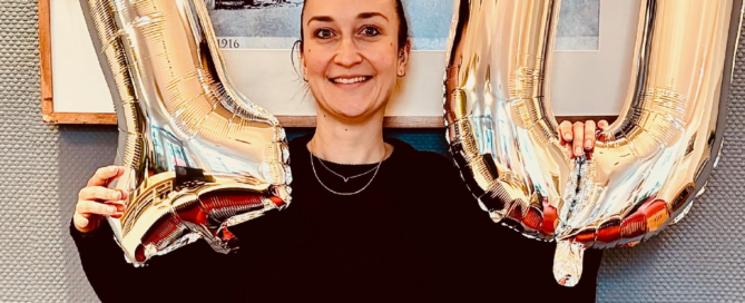 Carina Hauck feiert ihr Zehnjähriges Dienstjubiläum in der Zahnarztpraxis Susanne Rhiem mit zwei goldenen Zahlen-Luftballons in der Hand, einem Lachen auf dem Gesicht und steht dabei vor einem Bild mit dem alten Hauptbahnhof in Eschweiler darauf.